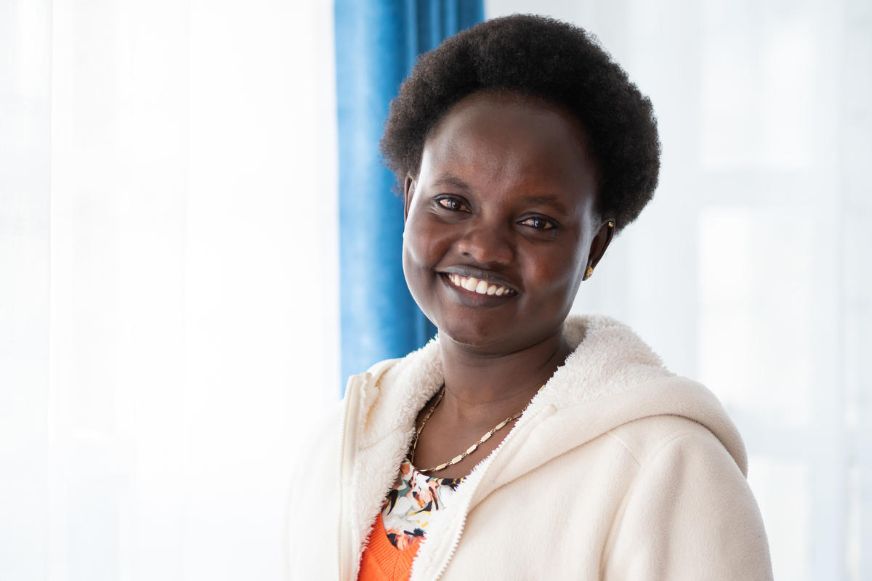 A smiling woman in a cream-coloured jacket stands indoors, facing the camera, with a blue curtain and bright background behind her. (Kenya, 2018)
