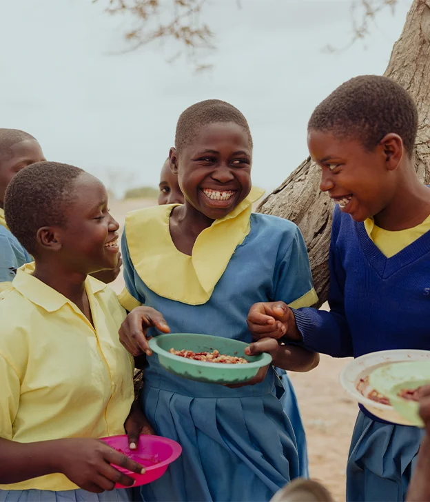 A group of students in school uniforms stand together outdoors, holding bowls of food under a tree.