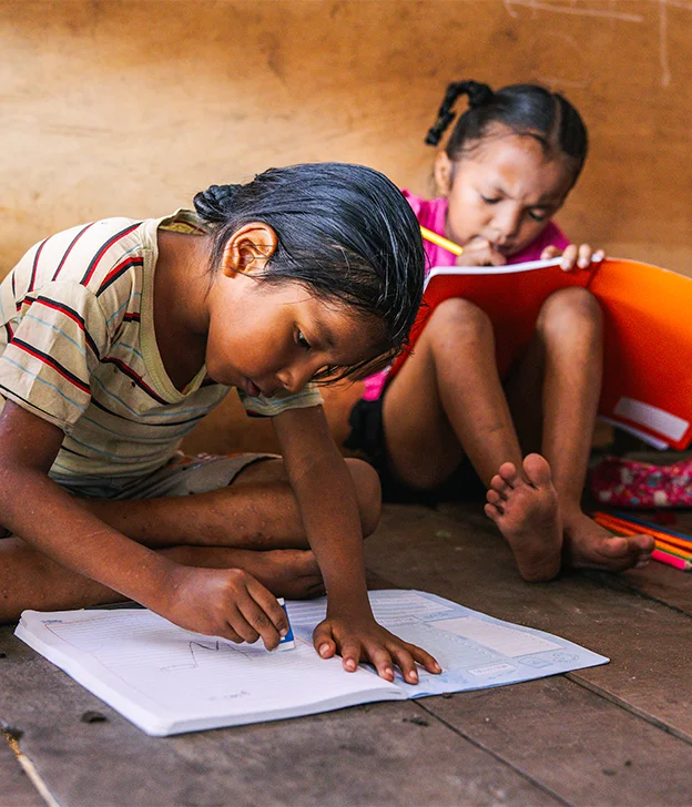 Two children sit on a wooden floor drawing and writing in notebooks with pencils and crayons inside a simple learning space.