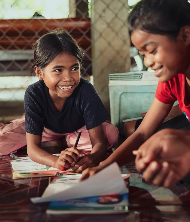 Children sit on the floor together, drawing and writing during a group learning activity indoors.