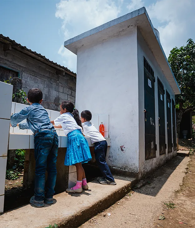 Three children are washing their hands at an outdoor sink.