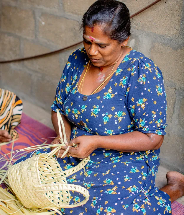 A woman sits indoors weaving a basket from palm leaves.
