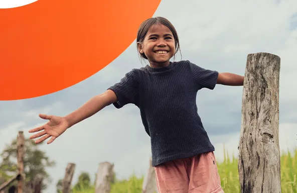 A child stands with arms outstretched beside a wooden post, smiling in a grassy rural field.