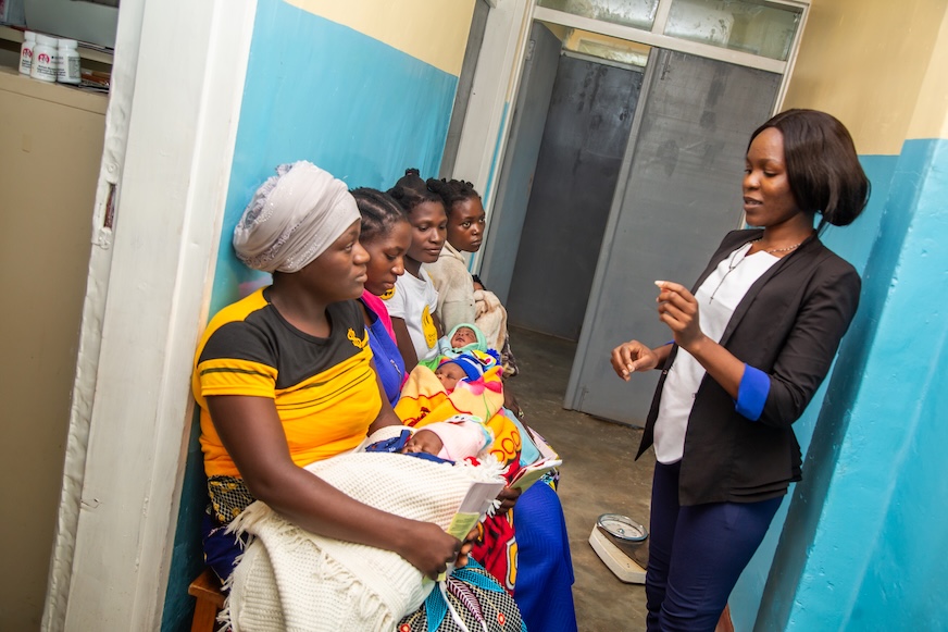 Four mothers sit on a bench with infants in their arms, while a woman in a blazer instructs them.