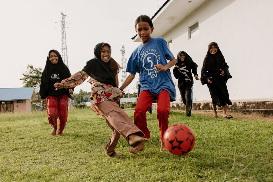 A group of girls laughs while kicking around a soccer ball. Some of the girls wear headscarves.