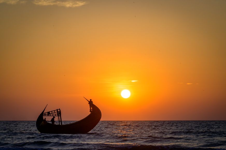 A Bangladeshi fishing boat at sea creates a striking silhouette against a vibrant orange sunset.