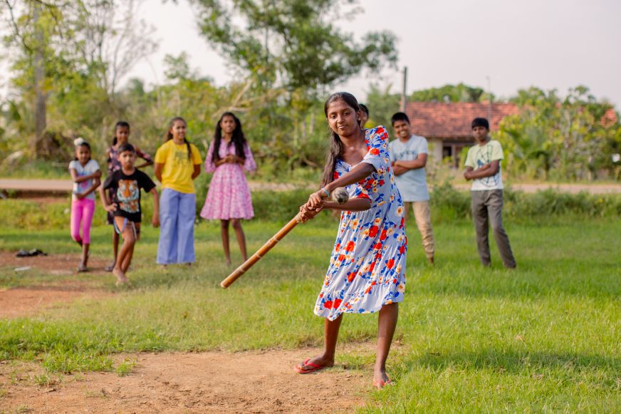 In Sri Lanka, an adolescent girl swings a makeshift cricket bat while other young people watch from the sidelines.