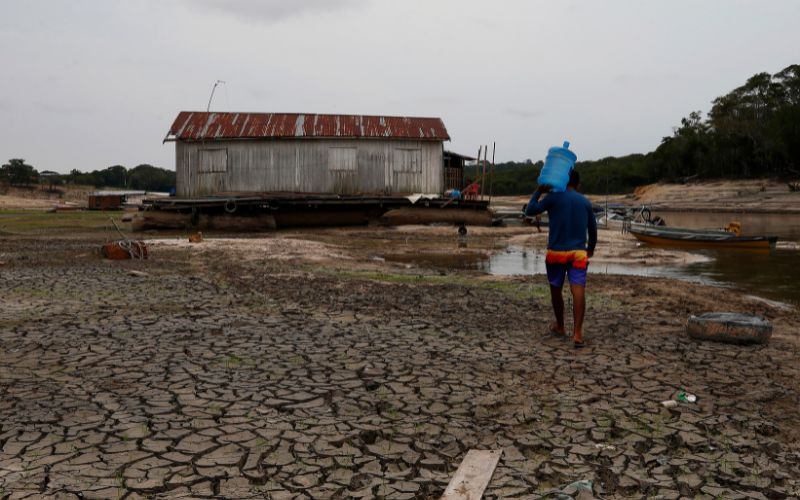A man walks across dried ground, carrying a water bottle on his shoulder.