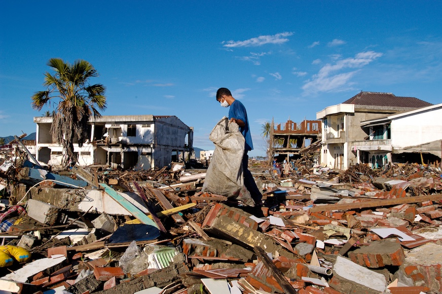A man wearing a medical mask looks through extensive rubble in a tropical setting.