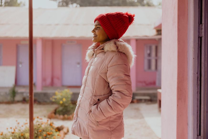 Profile of a smiling girl wearing a pink coat and red toque with a pink building in the background. (Nepal, 2024)