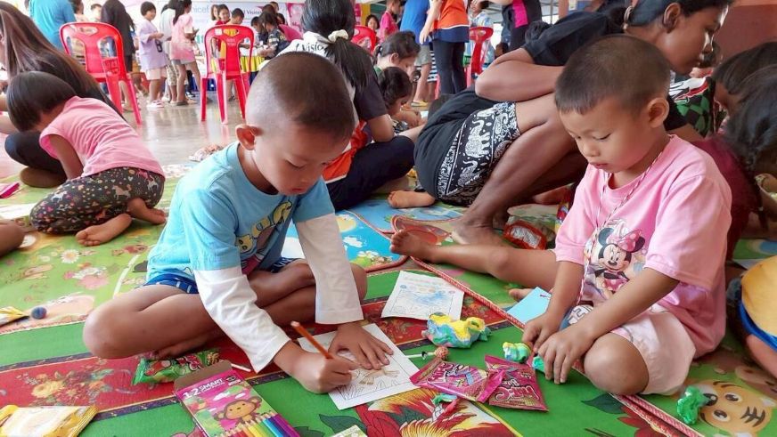 Two young boys sit cross-legged on a green mat, drawing pictures. (Thailand, 2025)
