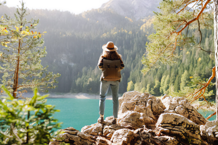 Woman standing on rocks wearing a backpack and Tilly hat overlooking a peaceful lake surrounded by trees.