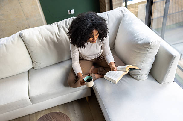 Woman sitting cross-legged on her couch reading a book with a cup of coffee