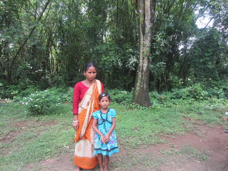 An adult and a child standing together in a rural, wooded setting.