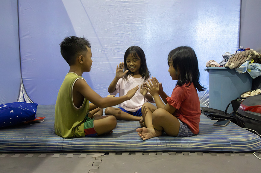 Three children sit in a temporary shelter, smiling and playing a clapping game.