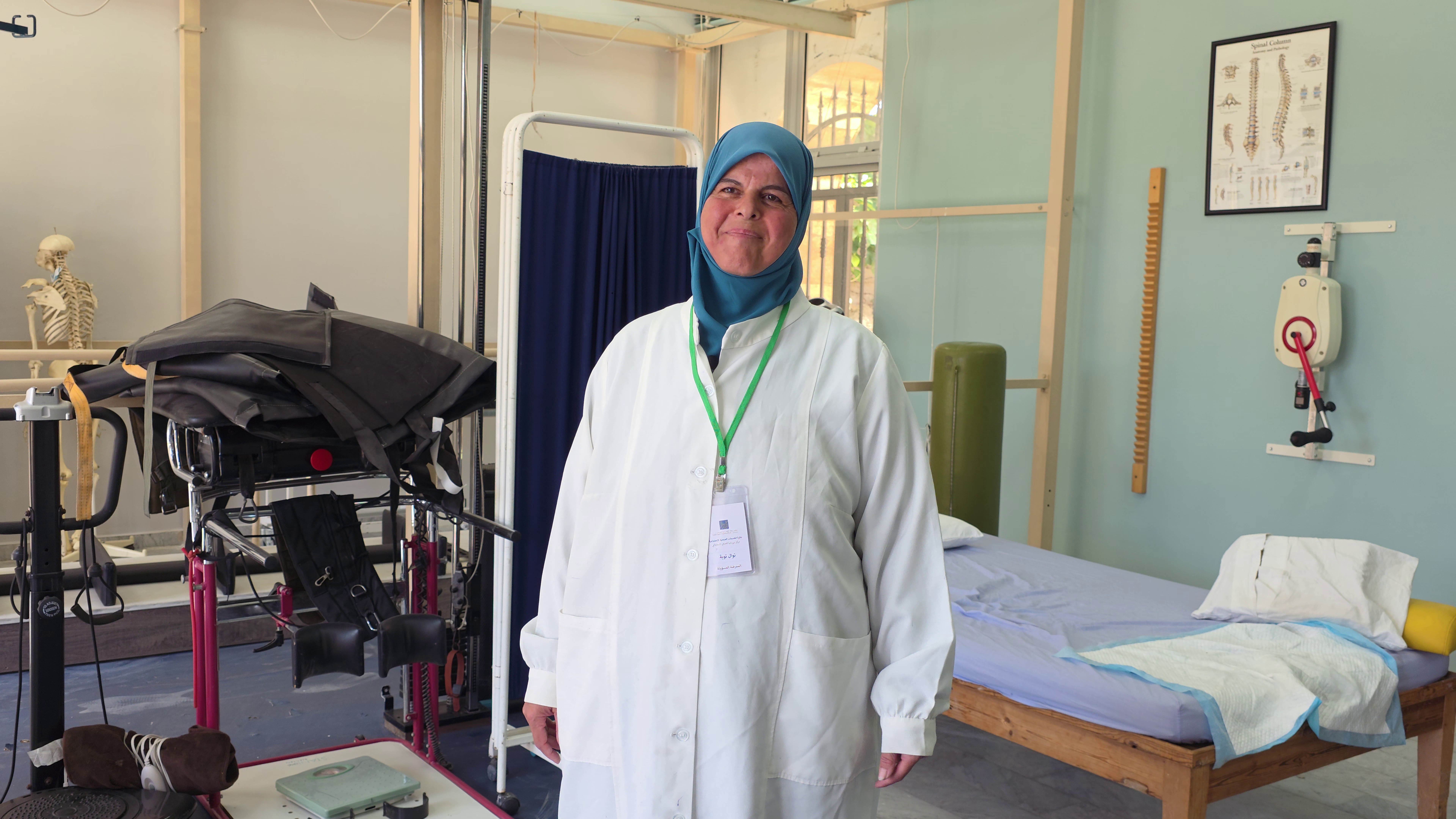 Nawal, a nurse, is wearing a lab coat and lanyard as she stands inside a room at the Derdghaya Socio-Medical Centre