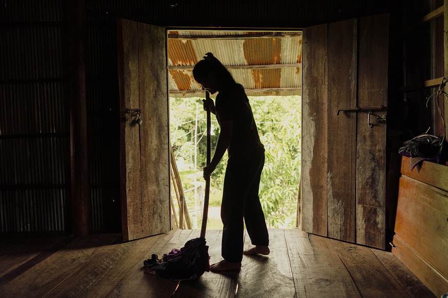 A silhouette of an adolescent girl sweeping the floor in a darkened room of her family’s hut in Cambodia.