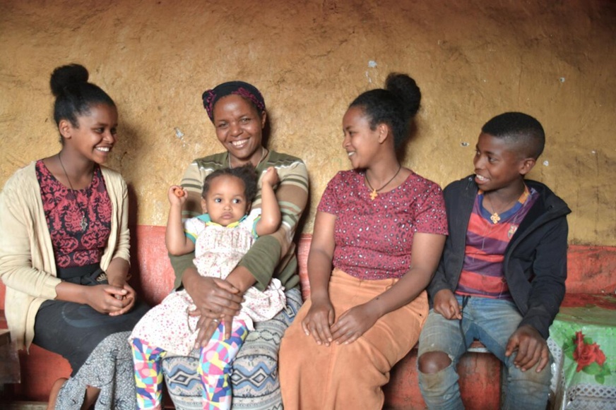 Five people sit closely on a bench against an earthen wall. A woman smiles brightly as she holds a small child on her lap while three young people of various ag