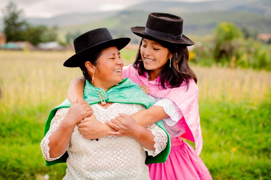 Two smiling females wearing wide-brimmed hats and bright, traditional-style clothing, stand in a grassy field, with the younger embracing the older one from beh