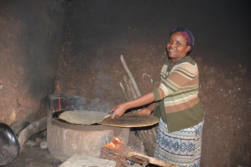 A smiling woman stands beside a wood-fired stove inside a rustic kitchen, using a flat utensil to cook flatbread over an open flame. (Ethiopia, 2025)