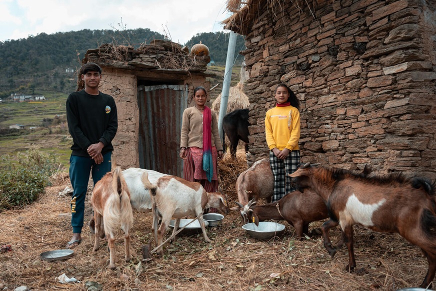 A mother and her two children stand in front of rustic stone buildings while several goats surround them eating from metal bowls on the ground. (Nepal, 2024)