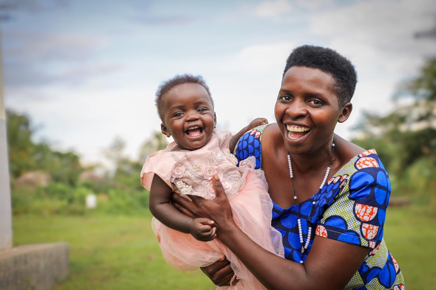 A smiling woman wearing a blue patterned top holds a laughing baby outdoors in a grassy setting. (Uganda, 2024)