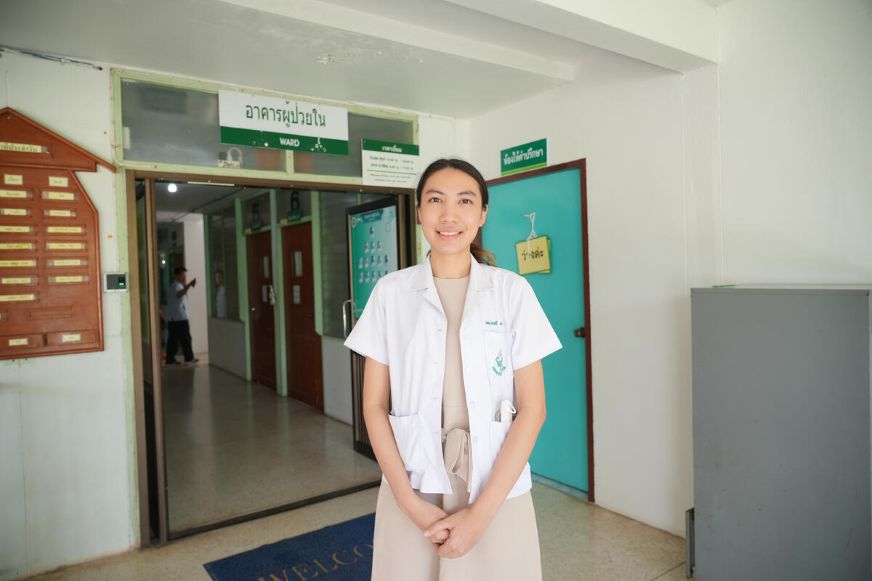 A female doctor wearing a white uniform stands in a brightly lit hallway, with doors and signage visible in the background, suggesting a clinic or hospital sett