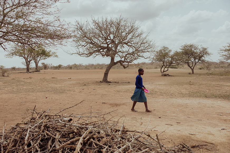 A girl carries an empty dish while walking on a dusty path in an arid landscape.