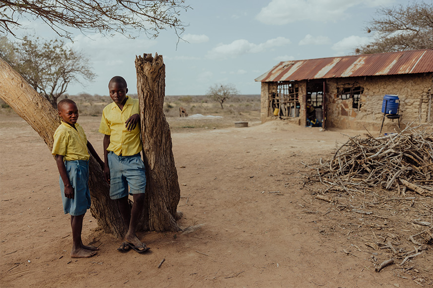 Two boys in school uniforms stand in a dry, dusty landscape, their run-down school in the background.