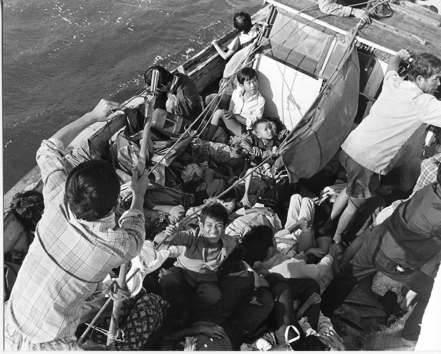 In a black and white image, children look up from a crowded boat at sea.