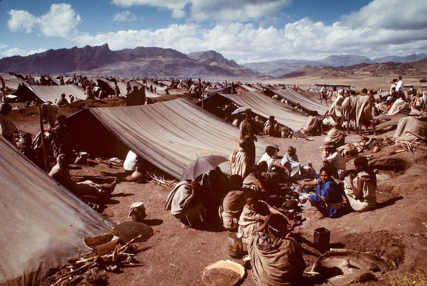 People sit amongst an expanse of makeshift tents in an arid landscape.