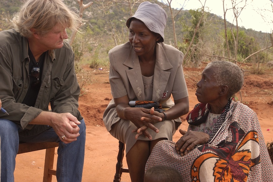 Two women sit speaking with a man outdoors.