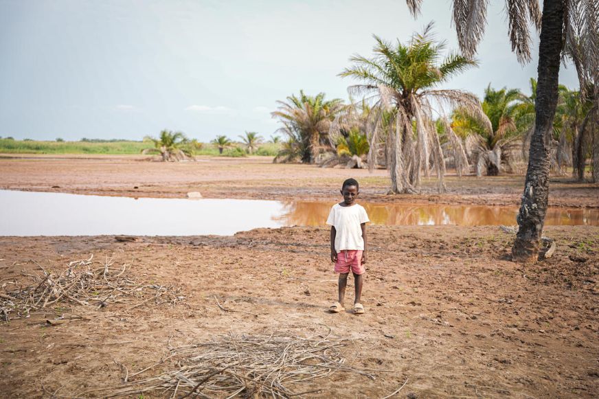 A child stands on dry, cracked ground near a shallow pool of water. Several palm trees grow behind the child, with sparse vegetation and open land extending int