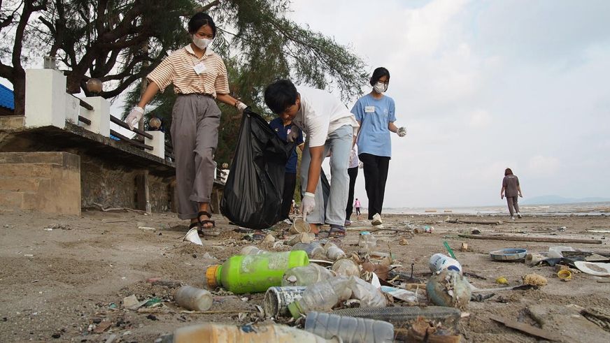 A group of young people wearing face masks collect plastic bottles and other trash from a sandy beach. One person holds a black garbage bag while others pick up