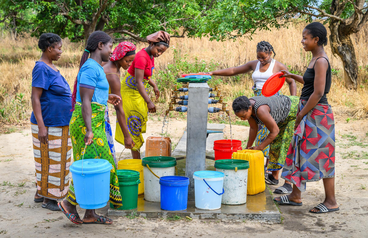 A group of people stand around a community water point in an outdoor setting, taking turns filling multiple plastic buckets and containers in various colours, w