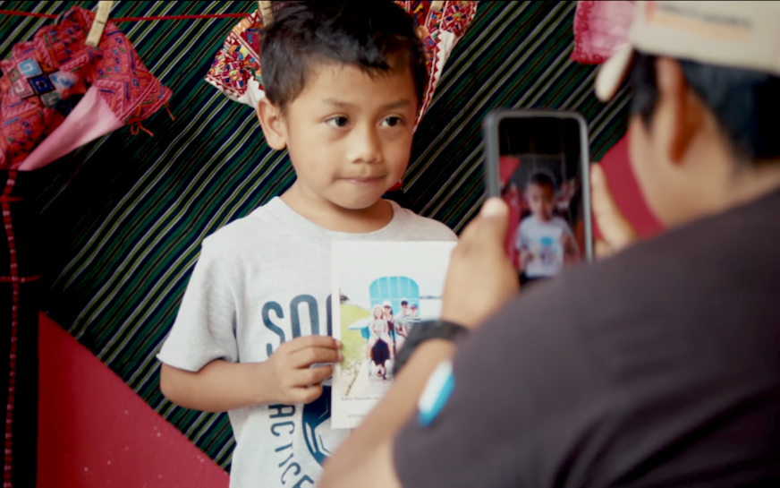 A little boy holds a picture of his sponsors while a staff member takes a photo on his phone.
