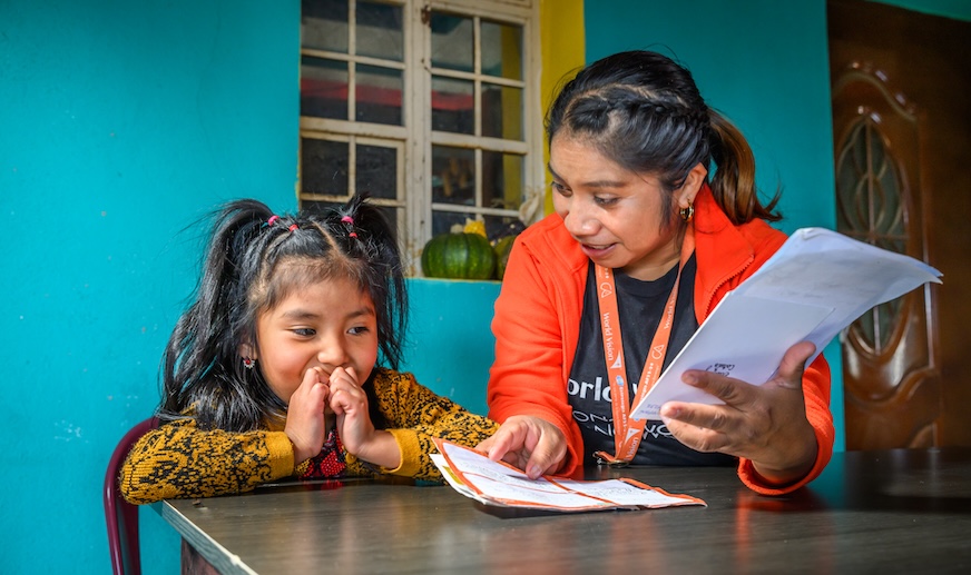 A young girl in Guatemala clasps her hands in front of her while a World Vision staff member shows her a letter.