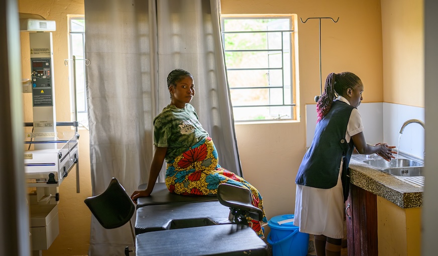 A pregnant woman sits on an exam table while a woman in an apron washes her hands in a stainless-steel sink.