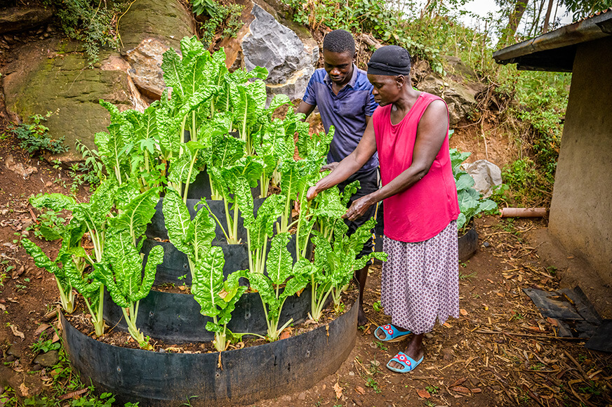 A man and a woman stand beside a circular, tiered “keyhole garden” made of dark recycled materials, filled with lush, vibrant green leafy vegetables growing in