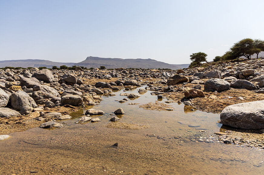 A rocky, dried-up riverbed under a clear sky, with only a few shallow, muddy puddles remaining among large boulders and distant hills.