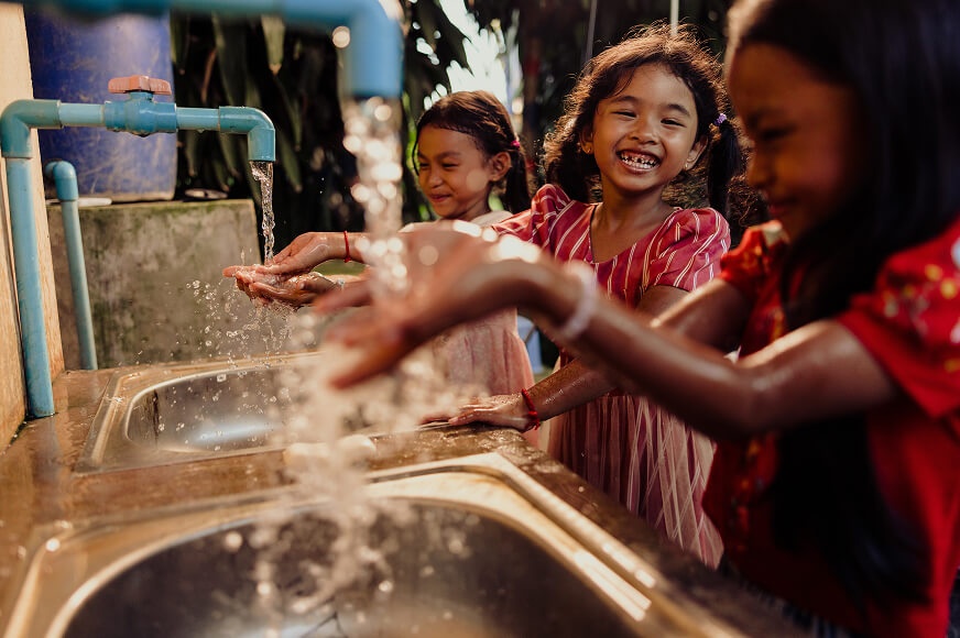 Three young girls laugh and smile while washing their hands under clean, running water at a row of outdoor sinks. The sunlight highlights the splashing water as