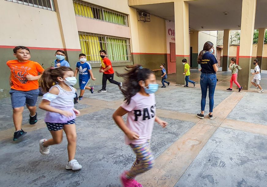 Children run around a large courtyard, wearing masks. A woman stands in the middle.