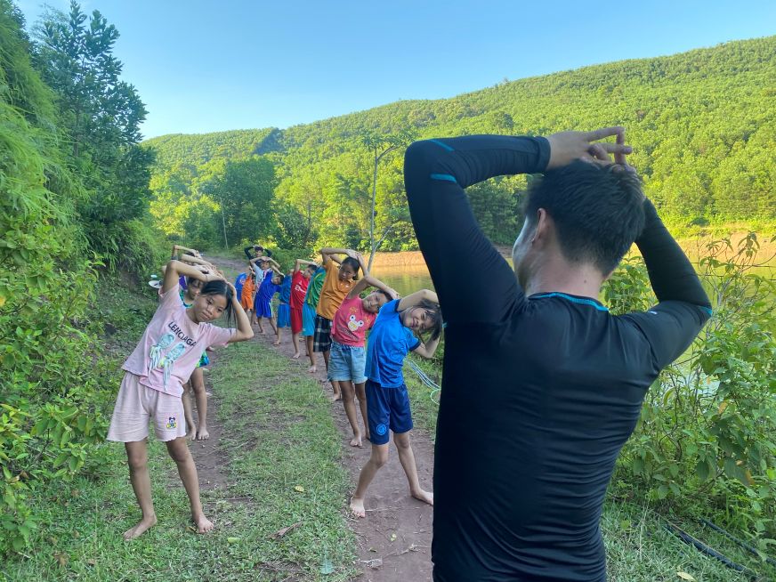 Two lines of children stretch to warm up, with a World Vision volunteer leading.
