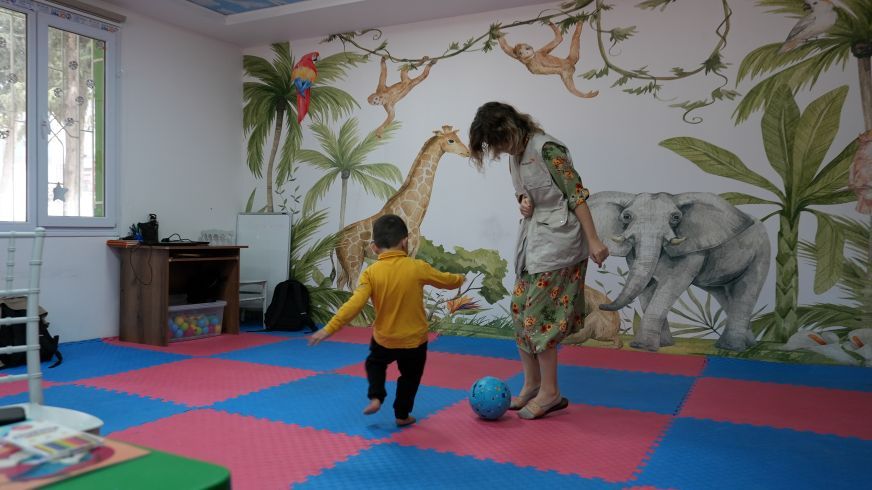 A woman and a small child kick a ball together inside a colourful indoor space for children.