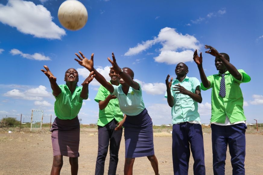 A group of male and female older adolescents stands stiffly in place while reaching for a ball that's coming their way.