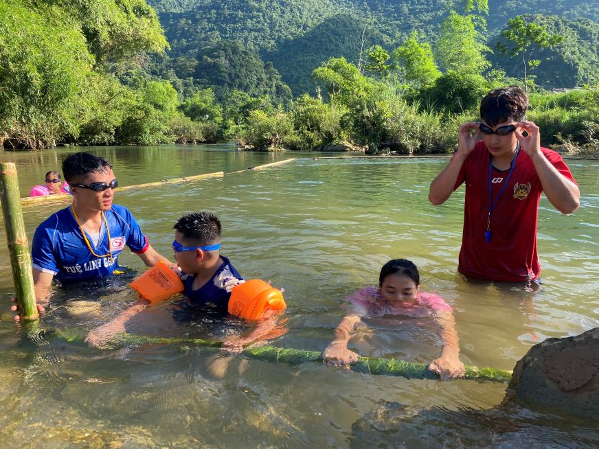 Two children grip a bamboo pole positioned horizontally in shallow water as two adults in goggles stand beside them.