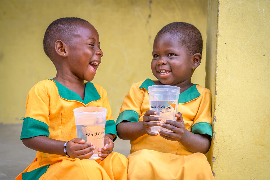 Two young children in yellow and green uniforms sitting together and laughing while holding clear cups of clean water. The cups feature the "World Vision Water"