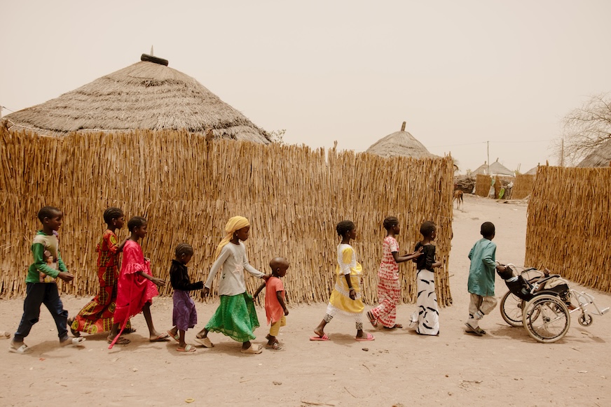 A young boy pushes a child in a wheelchair, while nine children follow in a line behind them.