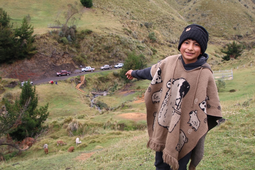 A young boy wearing a winter hat and knit poncho gestures to alpacas on the hill below him.