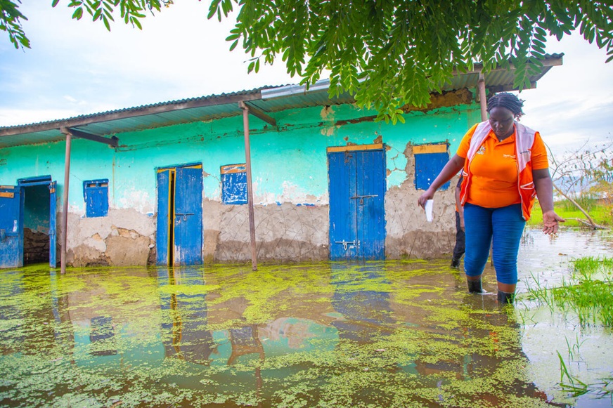 A woman wearing an orange World Vision uniform carefully walks through a flooded yard, past a green building with blue doors. (Uganda, 2024)